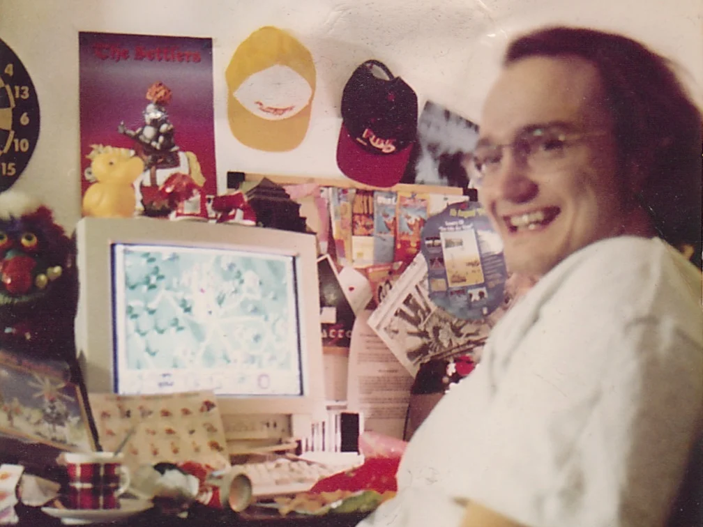 A photo print from the nineties: I sit at a crowded desk with long hair and glasses and laugh into the camera. On the tube monitor a snowy map from The Settlers II, on the wall a Settlers poster, two caps and a dartboard